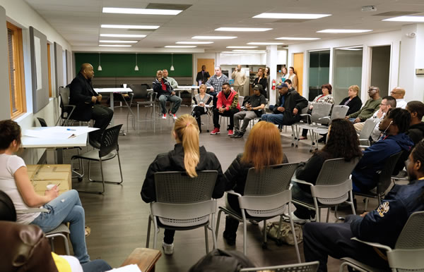 A room of people sitting in folding chairs, all listening to a man at the front of the room