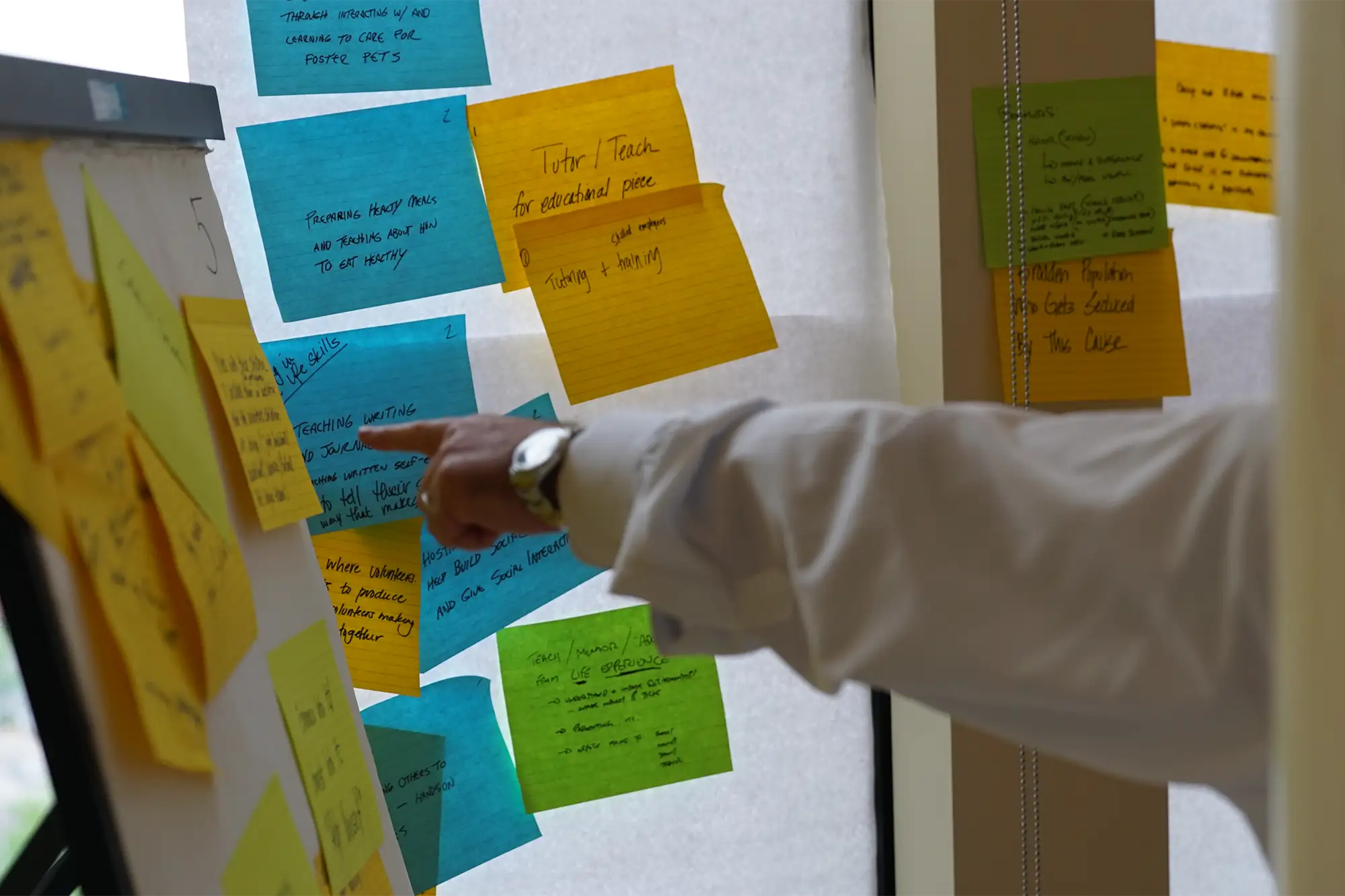 Someone pointing at a sticky note. There are multiple colored sticky notes all over the wall