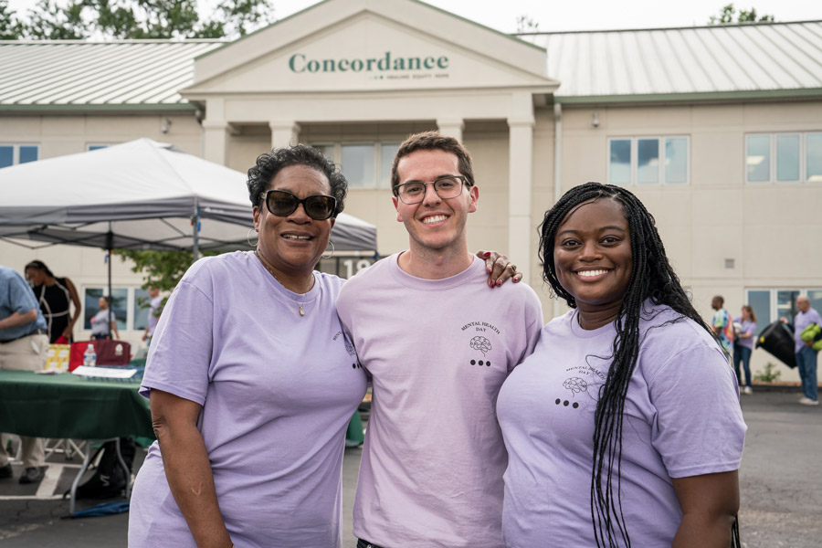 Three people in matching shirts stand in front of the concordance building and smile.