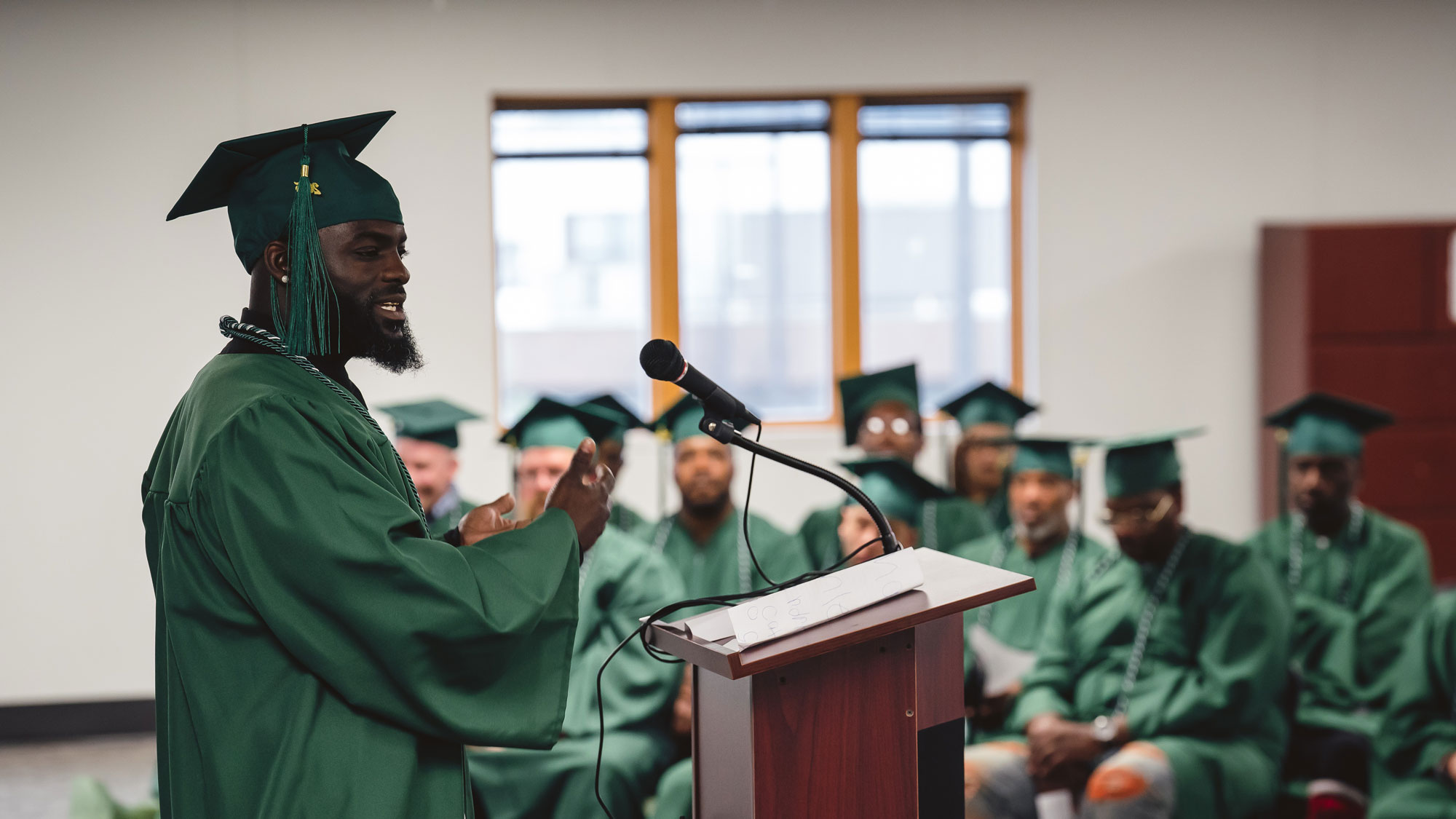 A commencement speaker giving a speech at a graduation. Fellow graduates seen in the background.