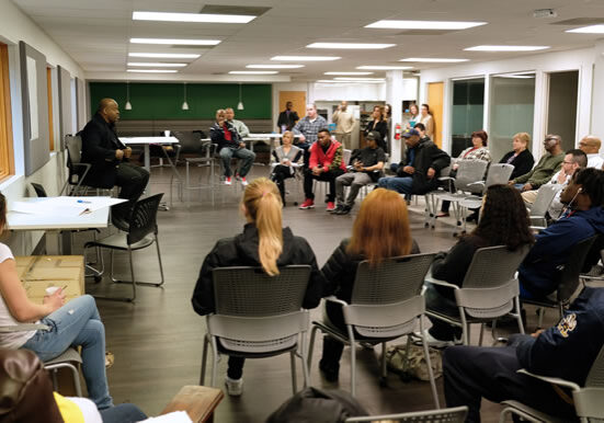 A room of people sitting in folding chairs, all listening to a man at the front of the room